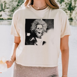 May include: A white cropped t-shirt with a black and white image of a skeleton with a large afro. The skeleton is smiling and has a skull face. The t-shirt is being worn by a person standing in front of a body of water.