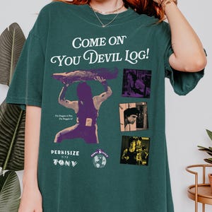 May include: Blue spruce-colored t-shirt with the text "Come On You Devil Log!" and a graphic of a person lifting a log. The shirt also features the text "Perkisize with Tony" and several small images. The bottom right corner of the shirt has the text "Blue Spruce".