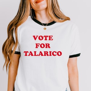 May include: A white ringer t-shirt with black trim on the collar and sleeves. The front of the shirt features the text VOTE FOR TALARICO printed in bold red capital letters. The shirt is paired with blue denim jeans.