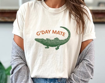 May include: Cream-coloured t-shirt featuring the text "G'DAY MATE" above a green alligator illustration. The shirt has short sleeves and a relaxed fit. The design is printed in a distressed style.