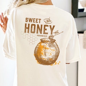 以下が含まれることがあります： 白いTシャツに、茶色のハチミツの壺とハチのグラフィックがプリントされています。Tシャツには「Sweet Like Honey」と「Proverbs 16:24」の文字が印刷されています。