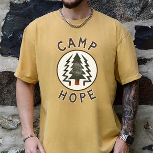 Peut inclure: Un t-shirt jaune moutarde avec un logo de style vintage qui dit "Camp Hope" avec un graphique d'arbre de pin à l'intérieur d'un cercle.