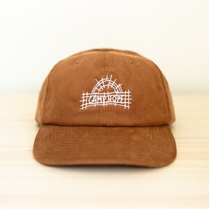 Peut inclure: Une casquette de baseball en velours côtelé marron avec un logo brodé blanc qui dit "Camp Hope" et un graphique de soleil.