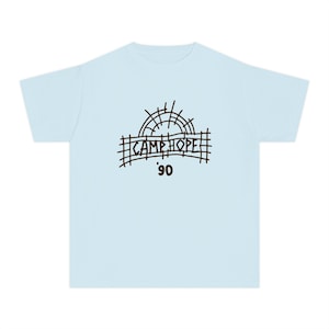 May include: Pale blue t-shirt with a black graphic. The graphic includes the words "CAMP HOPE" above the number "90" and a sun-like design. The shirt is made of a soft material.