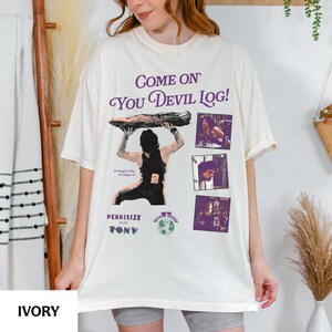 May include: Ivory-colored t-shirt with the text "Come On You Devil Log!" in purple script. The shirt features a graphic of a person lifting a log, with additional images and text. The shirt is a relaxed fit.