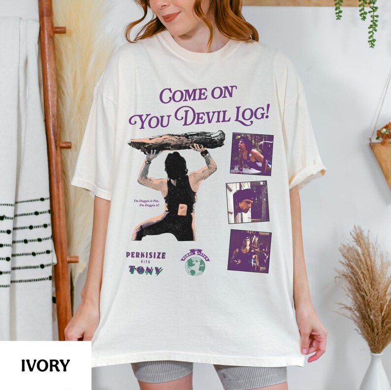 May include: Ivory-colored t-shirt with the text "Come On You Devil Log!" in purple script. The shirt features a graphic of a person lifting a log, with additional images and text. The shirt is a relaxed fit.