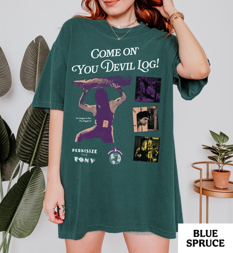 May include: Blue spruce-colored t-shirt with the text "Come On You Devil Log!" and a graphic of a person lifting a log. The shirt also features the text "Perkisize with Tony" and several small images. The bottom right corner of the shirt has the text "Blue Spruce".