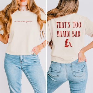 May include: A light beige t-shirt with the text "I'm tired of this Grandpa" on the front and "THAT'S TOO DAMN BAD" on the back. The shirt is being worn by a person with long brown hair and blue jeans.