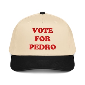 Puede incluir: Gorra de béisbol con corona color crema y visera negra. La gorra presenta el texto bordado "VOTE FOR PEDRO" en rojo, en referencia a una película popular. La gorra tiene una visera curva y un botón negro en la parte superior.
