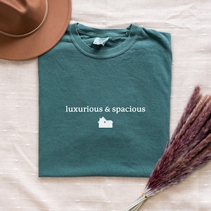 以下が含まれることがあります： 前面に「luxurious & spacious」という文字と小さな家のアイコンが白でプリントされた、ティールグリーンのTシャツです。