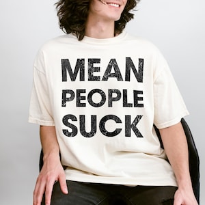 May include: Cream-colored t-shirt with the text "MEAN PEOPLE SUCK" in bold, black letters. The shirt has short sleeves and a crew neck. The text is centered on the front of the shirt.