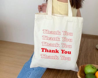 May include: A white canvas tote bag with red text that says "Thank You" repeated five times. The bag is being held by a person wearing a yellow tank top and blue jeans.