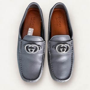 May include: A pair of grey leather loafers featuring a silver-toned metal GG logo hardware across the vamp. The interior is lined with brown leather, and the insole is stamped with the text GUCCI made in Italy in a gold-colored font.