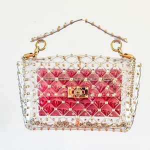 May include: A clear, studded handbag with a red interior. The bag features gold-colored hardware, including a chain strap and a clasp closure. The clear exterior is adorned with small, gold-colored studs.