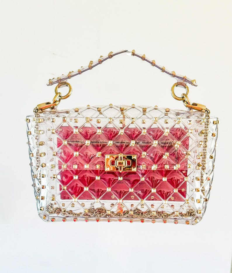 May include: A clear, studded handbag with a red interior. The bag features gold-colored hardware, including a chain strap and a clasp closure. The clear exterior is adorned with small, gold-colored studs.
