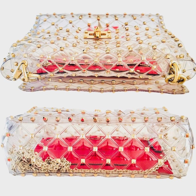 May include: A clear, transparent handbag with gold-colored studs and hardware. The bag has a red interior and a gold chain strap. The bag is rectangular with a flap closure and a gold clasp.