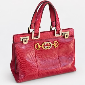 Könnte beinhalten: Eine rote Handtasche mit texturiertem Schlangenledermuster, ausgestattet mit zwei Tragegriffen und silberfarbenen Beschlägen. Die Vorderseite zeigt ein markantes gold- und silberfarbenes Metall-Trensenemblem mit dem ineinandergreifenden GG-Logo.