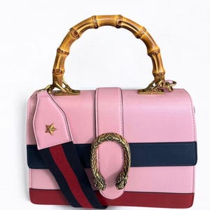Puede incluir: Bolso de mano de cuero rosa con asa de bambú y cierre metálico decorativo. El bolso presenta una franja azul marino y una base roja. También se incluye una correa para el hombro con una estrella y rayas rojas y azul marino.
