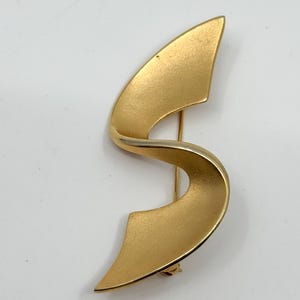 May include: A gold-toned brooch with a flowing, ribbon-like design. The brooch has a pin closure on the back and a matte finish. The shape is reminiscent of an abstract letter "S".
