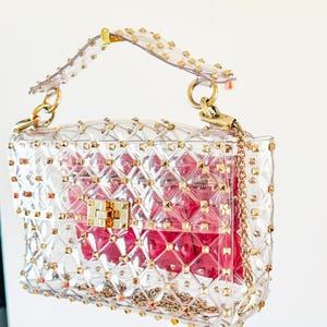 May include: Clear, studded handbag with gold-tone hardware. The transparent bag features a quilted pattern and a gold chain strap. A pink interior is visible through the clear material. The bag has a top handle and a clasp closure.
