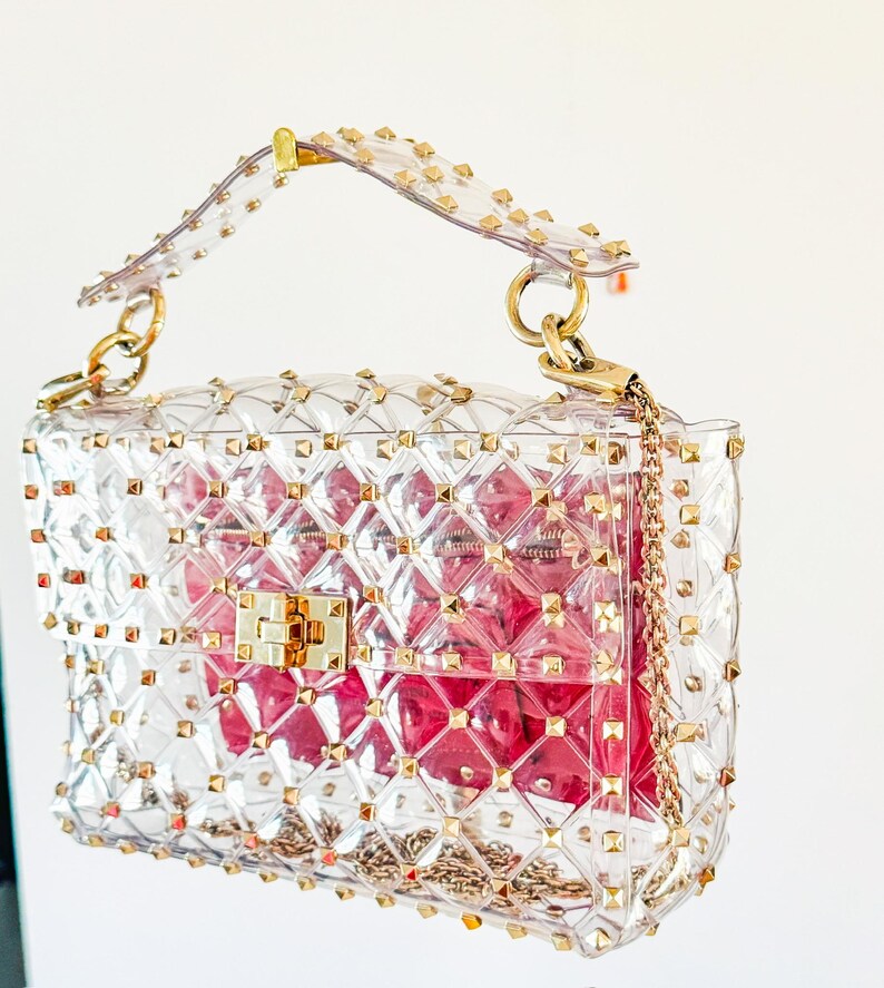 May include: Clear, studded handbag with gold-tone hardware. The transparent bag features a quilted pattern and a gold chain strap. A pink interior is visible through the clear material. The bag has a top handle and a clasp closure.