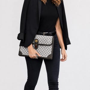 May include: A gray and brown patterned clutch with a gold-colored clasp and red tassel. The clutch is held in front of a person wearing a black blazer, black top, and black jeans. The clutch has a repeating pattern.