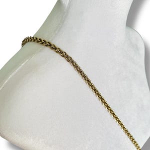 May include: A gold-colored chain necklace draped over a white mannequin. The chain has a textured, woven design. The background is a soft white, creating a contrast with the metallic chain.