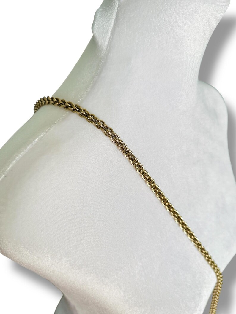 May include: A gold-colored chain necklace draped over a white mannequin. The chain has a textured, woven design. The background is a soft white, creating a contrast with the metallic chain.