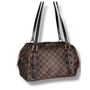 Puede incluir: Bolso de mano a cuadros marrón y beige con dos correas largas negras. El bolso tiene herrajes dorados y detalles de cuero. El bolso rectangular tiene cierre de cremallera y un pequeño bolsillo lateral.