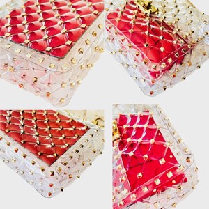 May include: Clear, transparent handbag with a red interior and gold-colored stud accents. The bag has a quilted pattern and a rectangular shape. The studs are evenly spaced around the edges of the bag.