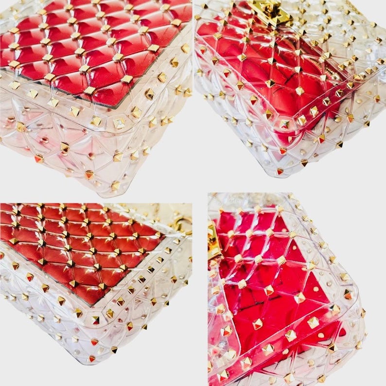 May include: Clear, transparent handbag with a red interior and gold-colored stud accents. The bag has a quilted pattern and a rectangular shape. The studs are evenly spaced around the edges of the bag.