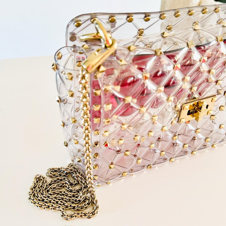May include: A clear, studded handbag with a gold chain strap and clasp. The bag has a quilted pattern and gold-colored studs. The interior of the bag is a reddish-pink color. The bag is sitting on a white surface.