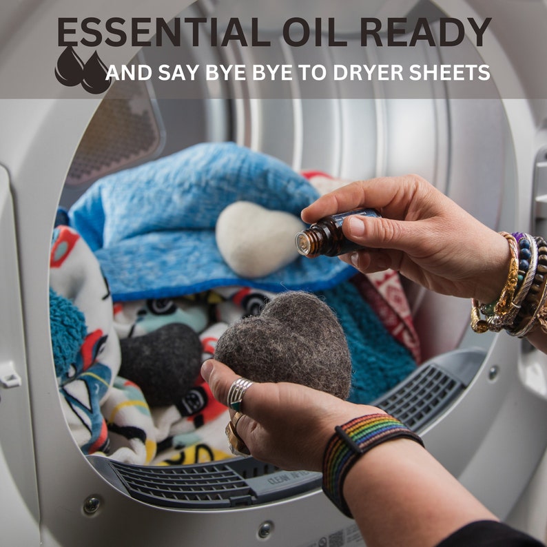 May include: A hand holding a gray heart-shaped wool dryer ball and a bottle of essential oil. The dryer ball is in a dryer with a blue blanket and other dryer balls.