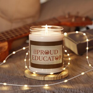 May include: A lit candle in a glass jar with the words "PROUD EDUCATOR" on a brown label. The candle sits on a round wooden coaster, surrounded by string lights. A guitar and a book are in the background.