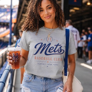 以下が含まれることがあります： グレーの半袖Tシャツで、青とオレンジの文字で「Mets BASEBALL CLUB QUEENS, NEW YORK」と書かれ、野球のグラフィックが描かれています。Tシャツはライトブルーのデニムショートパンツ、白いトートバッグ、ロゴ入りの透明なプラスチックカップと組み合わされています。