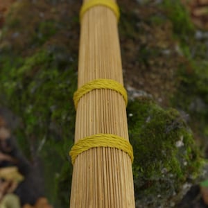 Peut inclure: Un faisceau cylindrique de fines tiges ou roseaux en bois brun clair, maintenu à intervalles réguliers par d'épaisses bandes de cordelette jaune tressée. L'objet est long et droit, avec une surface texturée créée par la disposition parallèle des éléments en bois.