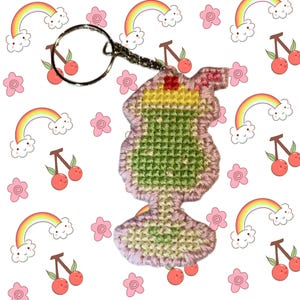 May include: A handmade keychain featuring a cocktail design. The keychain is crafted with a cross-stitch technique, showcasing a green, yellow, and pink cocktail with a cherry on top. The keychain is attached to a silver-colored metal ring.