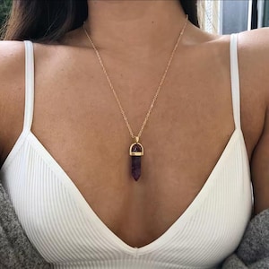 May include: A gold-colored chain necklace with a pendant featuring a purple crystal. The crystal is shaped like a pointed cylinder. The necklace is worn against a white top.