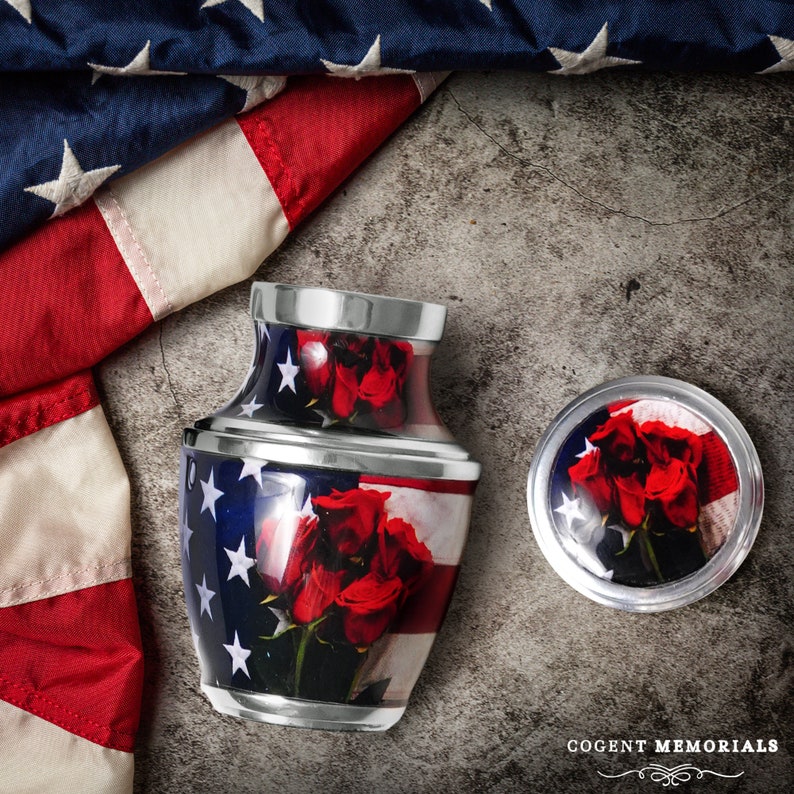 May include: A memorial urn with an American flag design, featuring red roses. The urn is accompanied by a matching lid, both with a silver trim. The background includes a folded American flag and a textured surface. The text "COGENT MEMORIALS" is visible.
