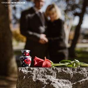 May include: A small, decorative urn with a red, white, and blue design featuring stars and flowers. A red rose and a few green leaves rest beside the urn on a stone surface. The words "COGENT MEMORIALS" are at the top.