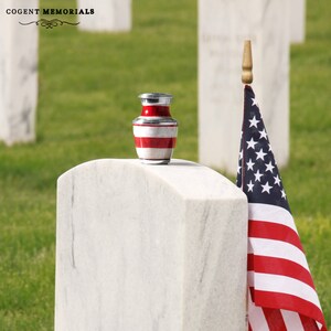 May include: A small, red, white, and silver memorial urn with horizontal stripes, resting on a white stone. An American flag is to the right. The text "COGENT MEMORIALS" is at the top left.