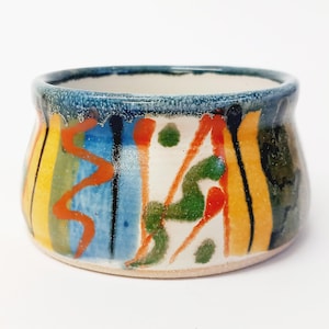 May include: A small, white ceramic bowl with a colorful, abstract design. The bowl has a wide, rounded base and a slightly flared rim. The design features stripes of yellow, orange, red, blue, and green, as well as black lines and dots.