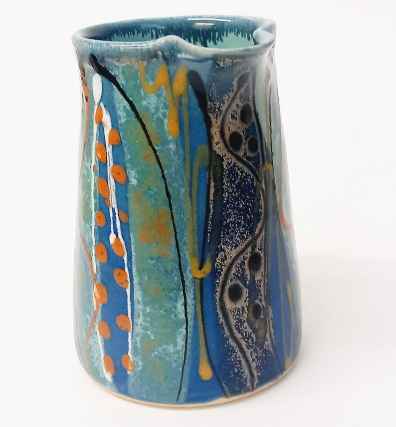A midnight blue jug is shown on a white background. The primary colour is blue with a turquiose oval and trailed lines in bright orange, yellow, white and black. The image shows the middle of the jug, the lip is pointing to the right.