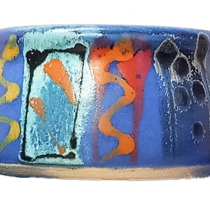 May include: A small, blue ceramic bowl with a colorful, abstract design. The bowl has a wavy, yellow line, a black outlined rectangle with orange and blue details, a red and orange wavy line, and a black and white abstract design.