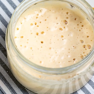 May include: A glass jar filled with a light beige, bubbly mixture. The jar is sitting on a white and gray striped fabric.