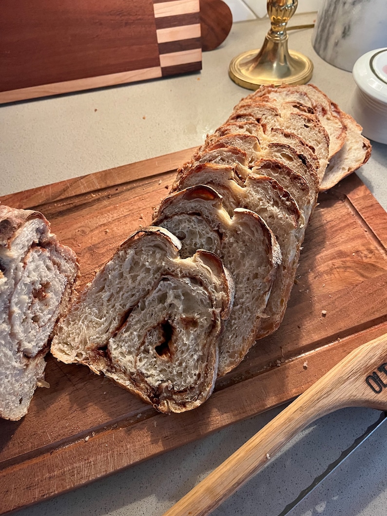 May include: A loaf of cinnamon swirl bread sliced on a wooden cutting board. The bread is light brown with a swirl of cinnamon throughout. The cutting board is made of wood and has a natural finish.