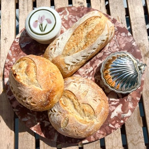 May include: Three loaves of artisan bread with decorative markings on a pink and brown marble surface. The bread is arranged in a circle with a small white ceramic dish with a floral design in the center.