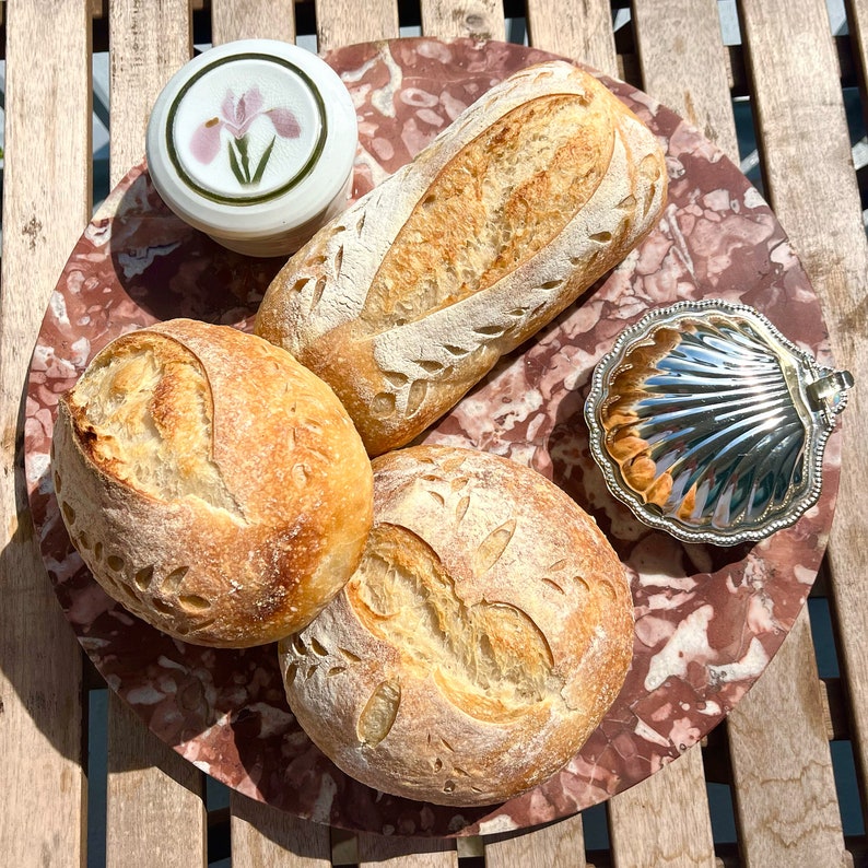 May include: Three loaves of artisan bread with decorative markings on a pink and brown marble surface. The bread is arranged in a circle with a small white ceramic dish with a floral design in the center.