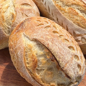May include: Three loaves of freshly baked sourdough bread with a decorative pattern on the top crust. The bread is golden brown and has a rustic appearance.