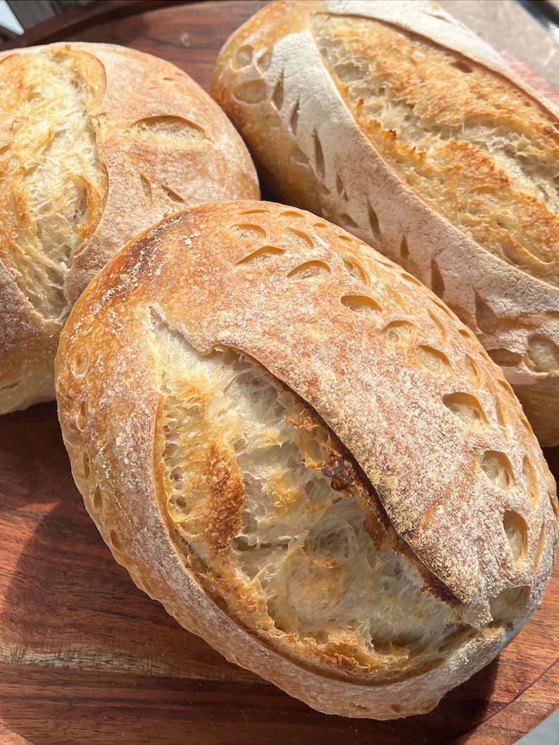 May include: Three loaves of freshly baked sourdough bread with a decorative pattern on the top crust. The bread is golden brown and has a rustic appearance.